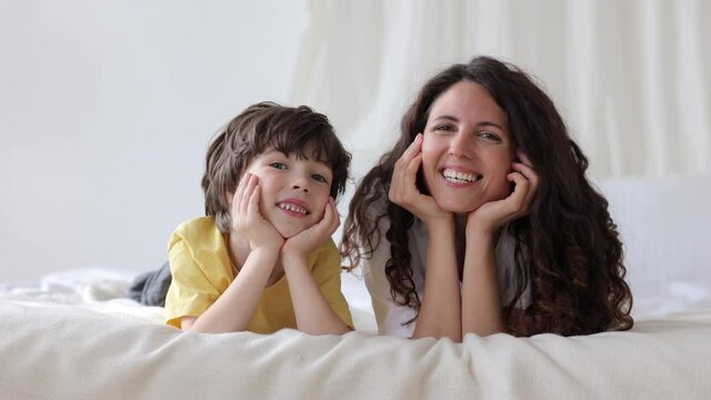 Cute family loving female parent and son lying on bed happy smiling. Morning portrait of young mother with cheerful little kid in bedroom. Curly caucasian girl, single mom or nanny with preschool boy