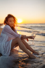 Fototapeta na wymiar Beautiful model in elegant white dress at sunset with glass of wine. The girl looks at the magical sunrise. Summer time. Travel, weekend, relax and lifestyle concept.