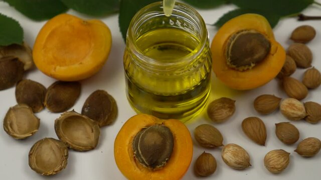 Drops of apricot kernel oil are dripped from a pipette into a small jar of oil. A small glass jar of the apricot kernel oil stands on a white table among the fresh apricots and the apricot kernels. Ap