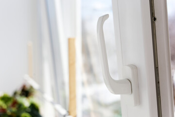 White handle is turned up on plastic window, which opened in ventilation mode for inflow of fresh air against background green leaves houseplants sill, daylight sunlight