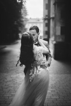 Stylish sensual bride and groom kissing on background of old church in rain. Provence wedding. Beautiful emotional wedding couple embracing. Romantic moment, black and white image