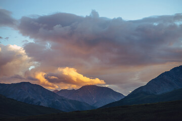 Beautiful mountain scenery with golden dawn light in cloudy sky. Scenic mountain landscape with illuminating color in sunset sky. Silhouettes of mountains on sunrise. Gold illuminating sunlight in sky