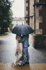 Stylish bride and groom kissing under umbrella on background of old church in rain. Provence wedding. Beautiful wedding couple embracing under black umbrella in rainy street. Romantic moment