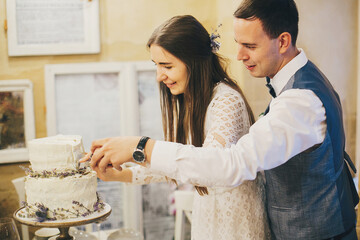 Stylish happy bride and groom cutting together modern cake with lavender on background of festive table in stylish restaurant. Provence wedding reception. Beautiful wedding couple celebrating