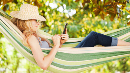 Smiling blonde woman with sunglasses using smartphone, lying relaxing on the hammock in the garden,...