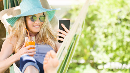 Smiling blonde woman with sunglasses using smartphone, relaxing on the hammock in garden, drinks a...
