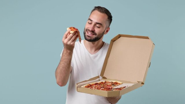 Smiling cheery charismatic joyful young brunet man 20s wears white t-shirt holding italian pizza in takeaway carton container box eating slowly with pleasure isolated on pastel light blue background.