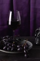 Red wine in a glass glass, grapes on a plate and part of a skull on a purple fabric background. The concept of magic, mysticism and esotericism.