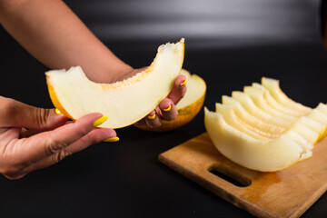 Female hand hold wedge of melon. Eating ripe yellow honeydew. Cut melon on a kitchen board.