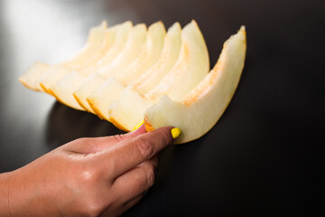 Hand picking piece of melon. Sliced honeydew on black table. Female hand holding tropical fruit.