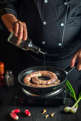 Professional chef adds oil to pan with raw homemade meat sausage. Preparation for cooking sausages in kitchen of a restaurant or cafe on table with vegetables.