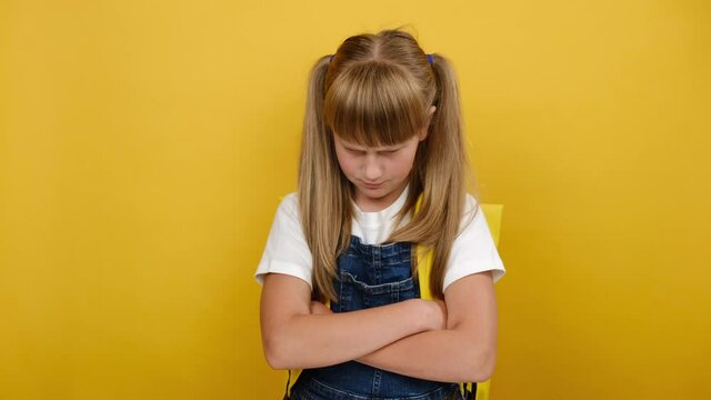 Portrait of angry sad little schoolgirl, crossed arms looking to side, does not want to go to school, wears backpack, posing isolated on yellow color background in studio. Children's emotions concept