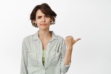 Skeptical young woman frowning, making disappointed smirk while pointing right, showing something upsetting, standing against white background