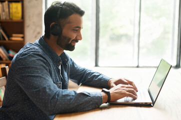 Young confident male Indian remote consultant wearing headset using laptop computer to virtual connection with customers or employees, customer service support manager talking at virtual meeting