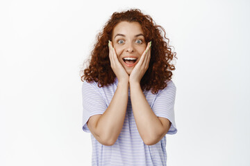 Surprised and excited redhead girl hold hands on face, staring in awe, impressed by smth cool, awesome news, standing in tshirt over white background