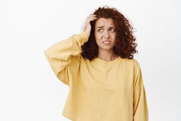 Fototapeta premium Confused awkward girl with curly hair, scratching head and looking aside clueless, dont understand, has no idea, puzzled, stands indecisive against white background