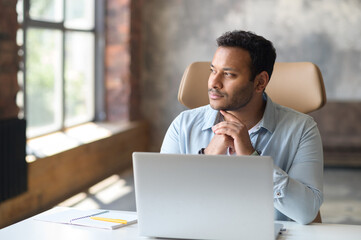 Serious concentrated clever indian freelancer guy using laptop for searching, looking away lost in...