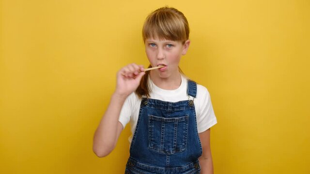 Portrait cute beautiful school girl kid with long blonde hair cleaning teeth with toothbrush, looking at camera, posing isolated over yellow color background wall in studio. Dental hygiene concept