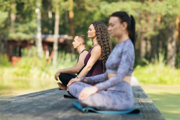 A trainer with two students doing meditation sitting on a mat in a lotus position on a wooden bridge in a park on a sunny summer morning