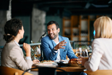 Happy entrepreneur talks to female co-workers while having business lunch at restaurant.