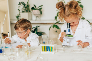 Boy and girl in white uniforms and protective glasses do chemical experiments in a home laboratory.Back to school concept.Young scientists.Natural sciences.Preschool and school education of children.