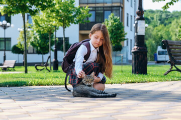 Cute little girl in school uniform and with backpack stroking a stray cat on the street, in the...