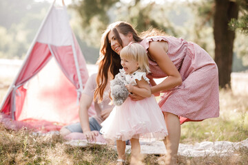 Mom, dad and little daughter are sitting next to wigwam decoration in the park. Family spending time outdoor in summer, having fun together. Girl are dressed in pink dress © Andriy Medvediuk