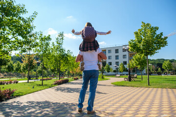 Naklejka premium Daughter in school uniform and with backpack sitting on her father's shoulders with outstretched arms. Dad giving daughter piggyback ride when going to school, rear view