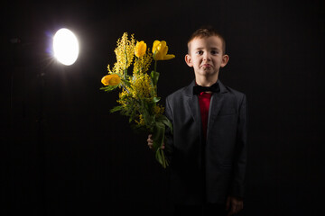 a boy with vitiligo in a red T-shirt, gray jacket and bow tie, with a bouquet of mimosa and yellow tulips, on a black studio background
