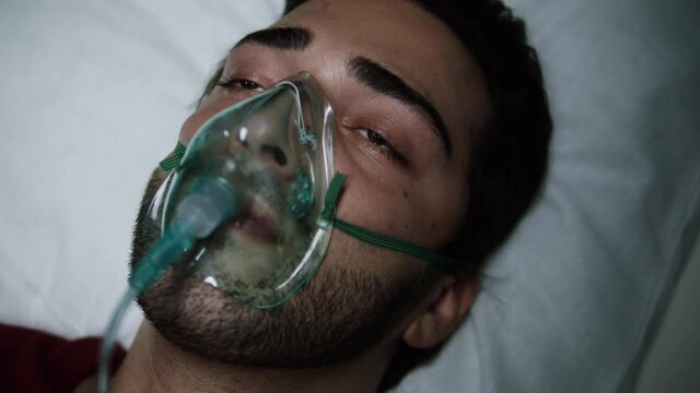 Patient with oxygen mask opening eyes after operation, lying in reanimation, bad human condition with coronavirus. Suffering covid man wearing oral-nasal mask with oxygen flow. Anesthesia face mask