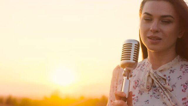 beautiful woman in white dress on top of mountain, on edge of abyss and sings song into shiny retro vintage microphone. Attractive lady singer on background of sunset or sunrise. Sun shine in camera.