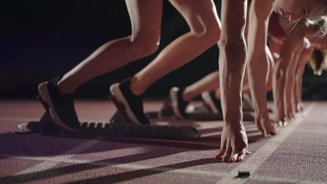 A row of runners womens crouch in the starting position before beginning to race. Females start with running shoes on the stadium from the start line in the dark with spotlights in slow motion.