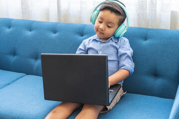 little latin boy with headphones using a laptop sitting on a blue sofa