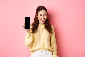 Image of cheeky attractive girl, showing you smartphone screen, winking and smiling, recommending...