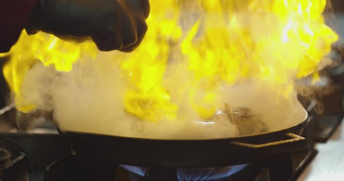 Professional chef cook flambe style . Prepares dish meat in a pan with open flames . Chef frying food on gas hob in commercial kitchen . Man frying steak in flaming pan on hob in restaurant .  