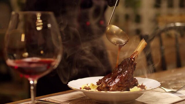 Spoon pouring sauce on meat. Cooked lamb knuckle . Dark red sauce poured on meat of lamb in plate with salad . Chef's Table with glass of wine and cooking show inspired footage .  Slow motion .