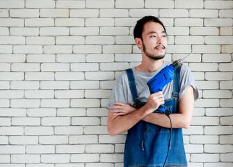 Young Asian man holding power drill standing in front of white brick wall, smiling and looking at camera