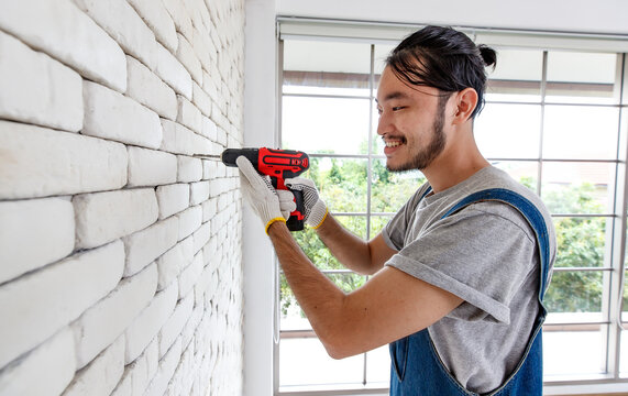 Young Asian man using electric drill on white brick wall in room, concept for home improvment, DIY work, do it youself