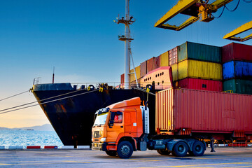 Truck carrying forty-foot container leaving port terminal with ship and quay crane on the...