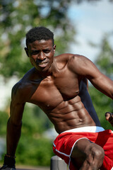 Muscular young man exercising at the sports field. African man looking to the side while doing horizontal bar exercises. Shirtless male model exercising outdoors.