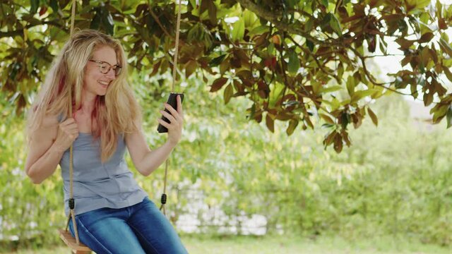 Smiling blonde woman with eyeglasses using smartphone, sitting on the swing in the garden, free time concept to surf the internet or chat with friends using social media