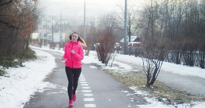 Running woman, headphone girl runner on the snow in winter sunny day. Female in pink sportswear jacket fitness training outdoors. Sport concept, leisure and freedom. 4K slow motion video 