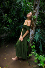 Caucasian woman leaning against a tree in tropical rain forest. Beautiful woman wearing long green dress. Walking trail in jungle. Nature concept. Travel to Asia. Bali, Indonesia