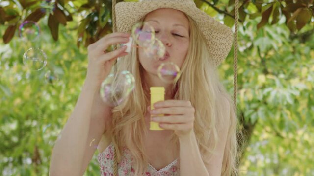 Smiling blonde woman wearing straw hat, makes soap bubbles sitting on the swing in the home garden, leisure and summer holiday concept