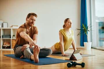 Fototapeta premium Young couple uses digital tablet while doing stretching exercises at home.