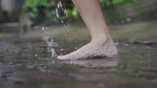 Low angle of female bare feet jumping and playing on water puddle on the raining day, pouring rain on the street side walking path way, fun time splashing water, jumping goof around, care free vibe