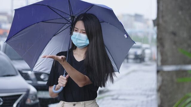 Young asian Woman wear face mask holding umbrella standing on the street side path, on the raining day, shower rainy season in Asia, car parked on the roadside, reach out her arm catching rain drops