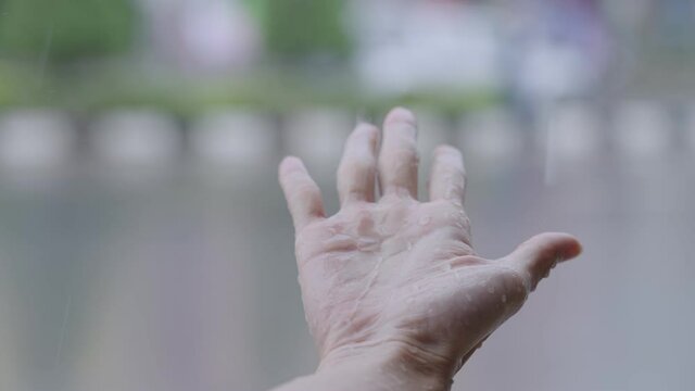 Female hand reaching out her hand to feel the rain drops, Under Heavy Rain Close Up. Catches rains on Palm, rainy season in slow motion, with urban street background, touch nature, wet hand from rain