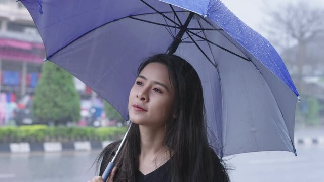 A young beautiful asian woman stands on roadside with holding a blue umbrella on rainy day caused by a tropical typhoon, waiting for bus on city roadside, weather forecasting, cold and flu prevention