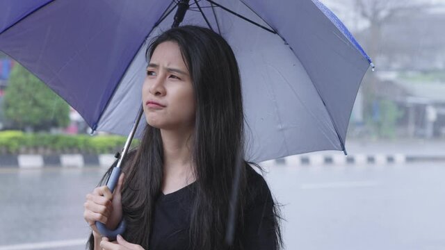 Young asian female with a long black hair disappointed stuck in heavy rain outside, hand using umbrella to prevent getting wet, facial expression of negative feeling, tropical climate changes